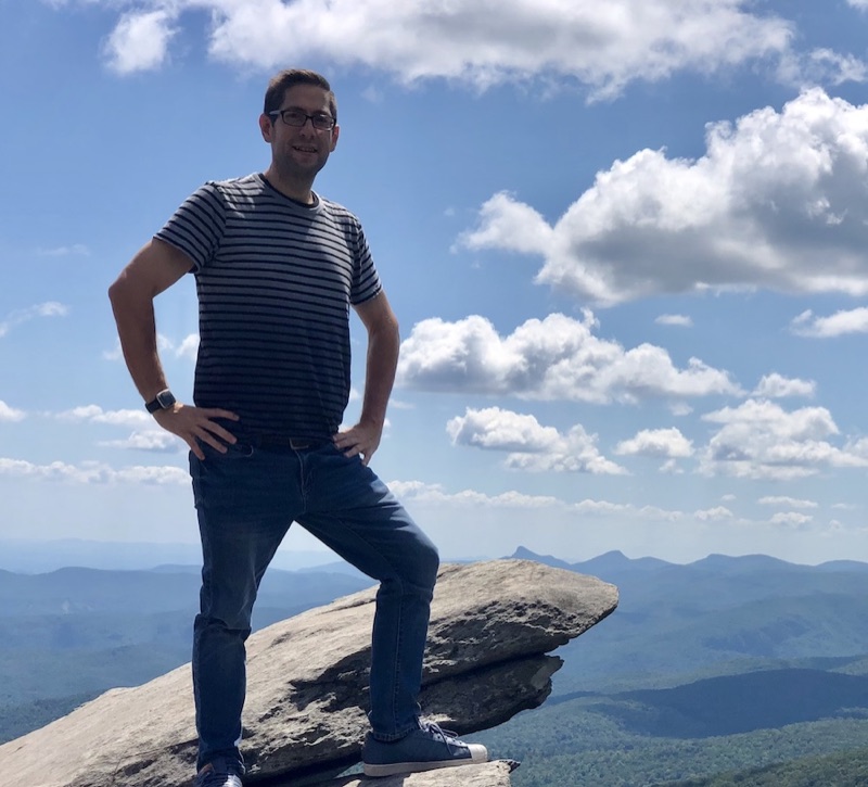 Kevin Young standing with one foot up on a rock ledge, with blue sky and mountains behind him